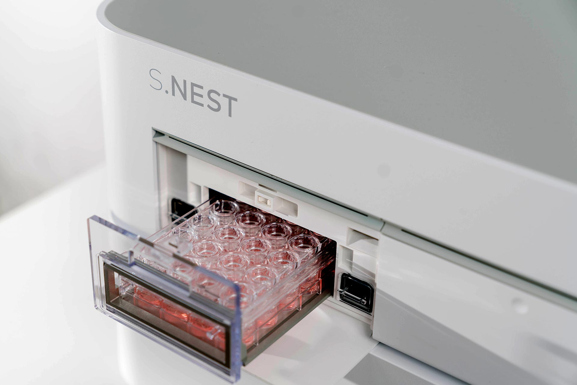 Ein weißes Laborgerät mit offener transparenter Schublade, in der ein Plastikbehälter mit vielen Löchern steckt, die mit einer rötlichen Flüssigkeit gefüllt sind.