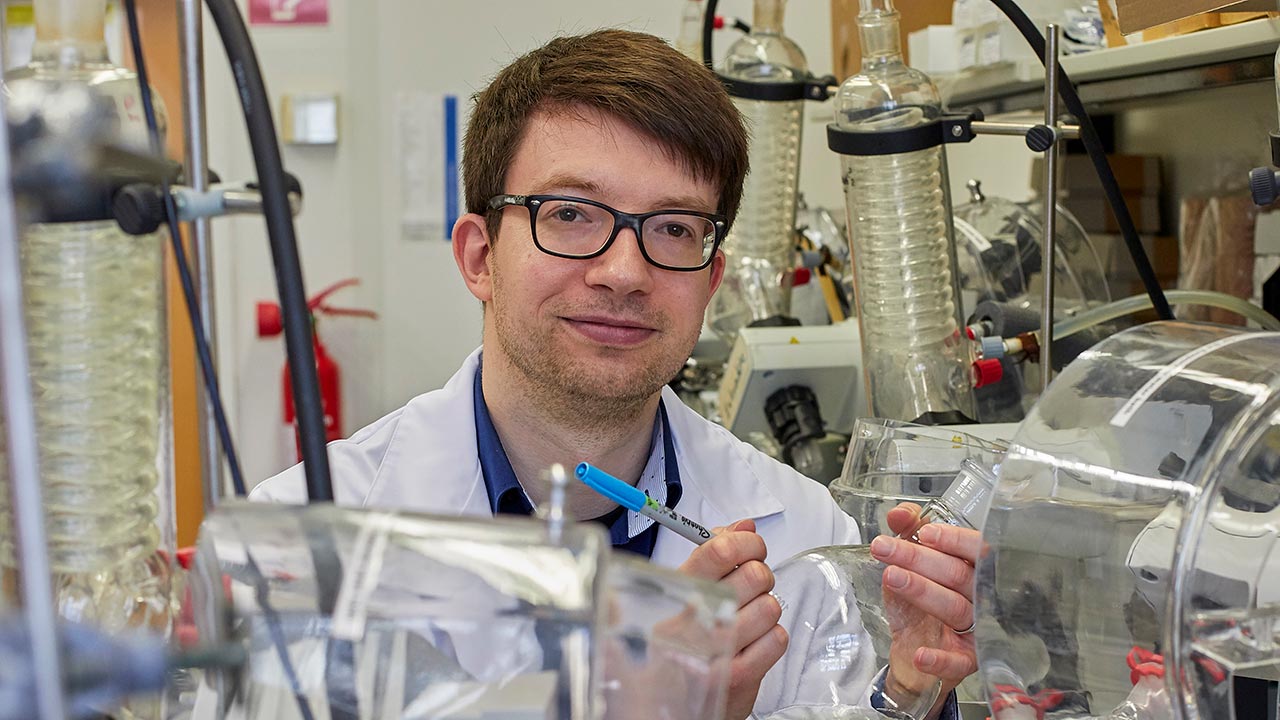 Ein junger Mann mit Brille und Laborkittel beschriftet im Labor einen Glaskolben