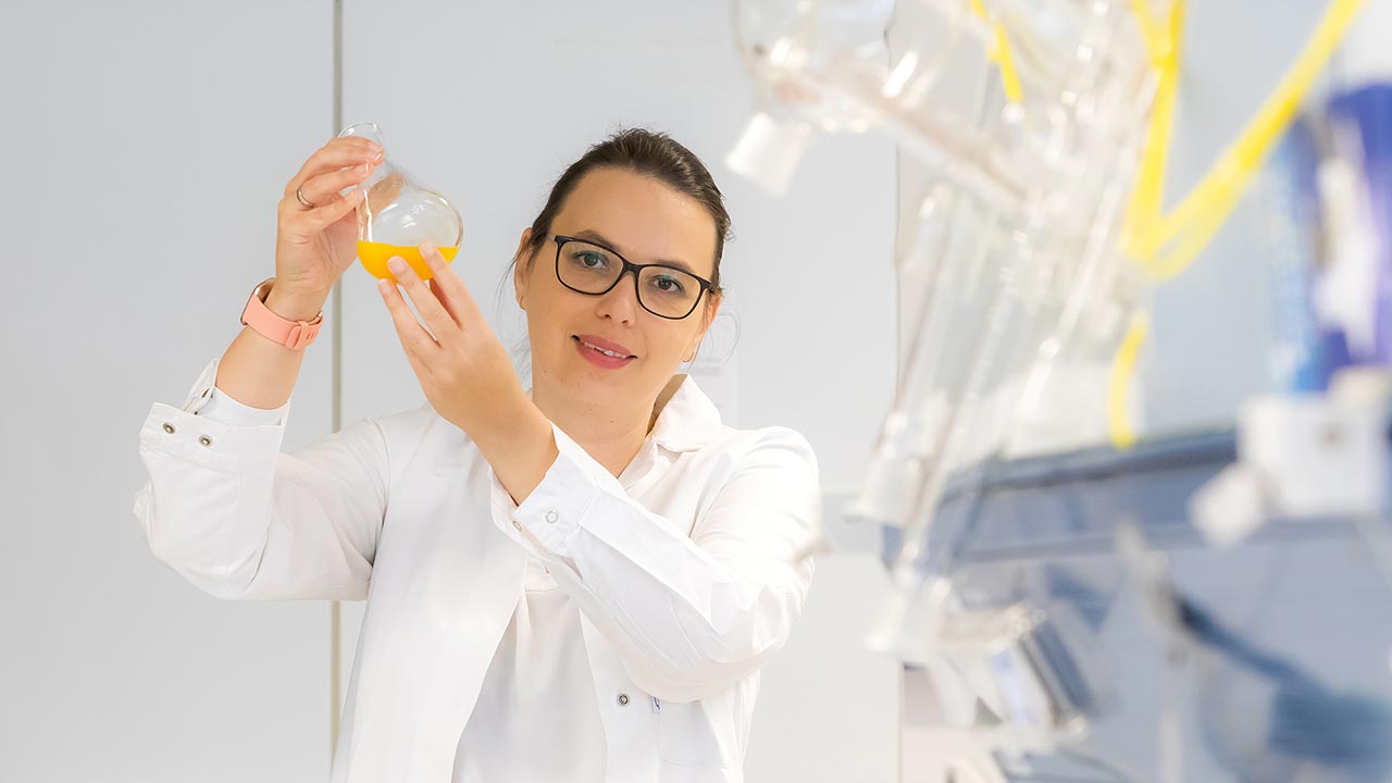 Eine Frau im Laborkittel hält einen Kolben mit gelber Flüssigkeit hoch.
