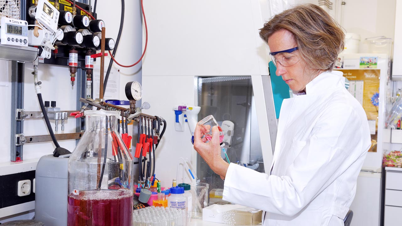 Frau im Laborkittel untersucht eine rötliche Flüssigkeitsprobe vor einer Laborbank.