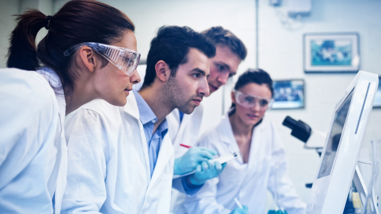 Ein Forscherteam gekleidet in weißen Laborkitteln schaut auf einen Monitor.